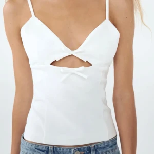 Vit croppad linnetopp med rosetter - Supersöt vit linnetopp från Zara med smala axelband och två små rosetter framtill. Toppen har en snygg cut-out detalj under bysten och är croppad i modellen. Perfekt att matcha med jeans eller kjol för en fräsch look.