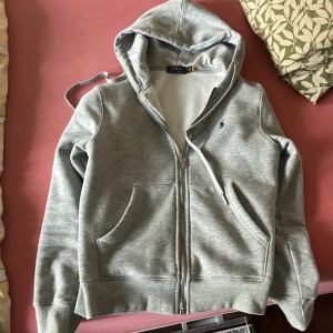 Grå hoodie från Polo Ralph Lauren - Snygg grå hoodie från Polo Ralph Lauren med dragkedja och huva. Tröjan har två stora fickor fram och ett broderat mörkblått Polo-logo på bröstet. Mjuk insida i fleece och ribbade muddar vid ärmslut och nederkant. Perfekt för chill eller skola.