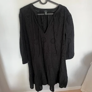 Svart broderad klänning från Lindex - Svart klänning från Lindex med v-ringning och knytband. Klänningen har broderade detaljer och hålmönster över hela tyget, samt trekvartsärmar. Tillverkad i bomull och har en luftig, avslappnad passform som ger en bohemisk vibe.