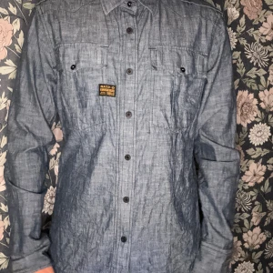 G-Star RAW Gould Chambray Shirt - Säljer en G-Star RAW Gould Chambray Shirt i klassisk militär/utility-stil. Färg: Mellanblå chambray (jeansliknande men tunnare tyg) Modell: Gould Chambray Shirt  Detaljer: Knappstängning med dold dragkedja under knappslån Två bröstfickor med knappar Epåletter på axlarna Liten G-Star RAW-patch på bröstet Passform: Regular fit, skjorta/overshirt-stil Material: 100% bomull (chambray)  Skjortan är perfekt som en lätt jacka eller overshirt, går att styla både avslappnat och mer🤗