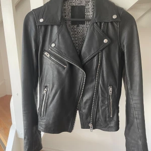 Svart skinnjacka från Rockandblue - Snygg svart skinnjacka från Rockandblue med coola dragkedjor och detaljer i silver. Jackan har klassisk bikerstil med sned dragkedja, flera fickor och spännen bak. Tillverkad i äkta skinn med foder i polyester. Perfekt för dig som vill ha en edgy look. Använd endast ett fåtal gånger, skick som ny.