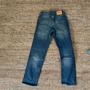 Blå jeans Jack & Jones - Snygga blå jeans från Jack & Jones, modell Regular Clark. Klassisk femficksdesign med orangea sömmar och läderpatch bak. Jeansen har en rak passform och passar då utmärkt till någon som har storlek 158-166.