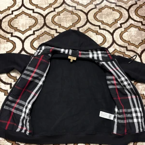 Svart Burberry hoodie med rutig insida - Svart hoodie från Burberry i storlek M med klassiskt rutigt foder i huvan och insidan. Tröjan har dragkedja, snörning i huvan och broderad Burberry-logga på bröstet. Tillverkad i mjuk bomull, perfekt för dig som gillar stilren streetwear med lyxig touch.