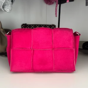 Rosa quiltad axelväska i mocka-look - Säljer en superfin axelväska i starkt rosa färg med quiltad design och bred axelrem. Väskan har en cool rutstruktur och är gjord i mjuk mocka-liknande material. Perfekt för dig som vill sticka ut med en färgglad accessoar.