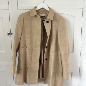 Beige kappa från Zara Basic XS - Stilren beige kappa från Zara Basic i storlek XS. Jackan har klassisk krage, stora fickor framtill och knäpps med svarta knappar. Materialet känns som en blandning av bomull och syntet, perfekt för vår och höst. Enkel och clean look som passar till allt.