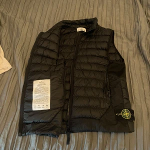 Svart dunväst från Stone Island M - Svart dunväst från Stone Island med ikonisk kompasspatch på sidan. Västen är quiltad, har dragkedja framtill och är tillverkad i lätt nylon med dunfyllning. Perfekt för lager-på-lager och streetwear-vibe. Skriv om du har frågor! Inte använd ofta fortfarande i top skick.!