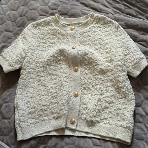 Beige tröja/kofta från H&M - Kortärmad krämvit kofta från HM. Storlek M- aldrig använd, helt i nyskick! Fint strukturstickning med eleganta guld knappar. Lätt att styla både till vardags med jeans eller över en klänning för en mer uppkläd  look! 