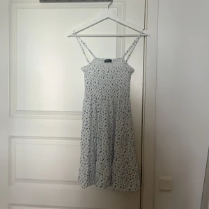 Vit blommig sommarklänning FB Sister - Supersöt vit klänning från FB Sister med blått blommönster och smock upptill. Klänningen har tunna axelband och en luftig, kort modell som passar perfekt för varma dagar. Den är lätt och flowy, vilket gör den riktigt bekväm och snygg.