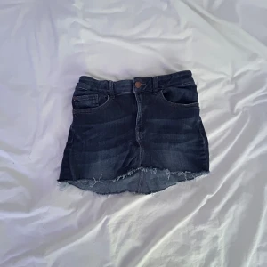 Mörk jeanskjol från Denim by Lindex - Snygg mörkblå jeanskjol från Denim by Lindex i modell slim. Kjolen har klassiska fickor fram och bak, rå kant nertill och knappstängning. Perfekt för dig som gillar en enkel men cool stil.