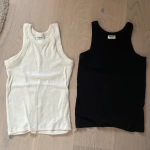 2-pack ribbade linnetoppar Kappahl - Två klassiska linnetoppar från Kappahl i storlek 158/164. Ena toppen är vit och den andra svart, båda i ribbad bomull med ärmlös design och rund hals. Perfekta att styla med jeans eller shorts för en clean och enkel look.