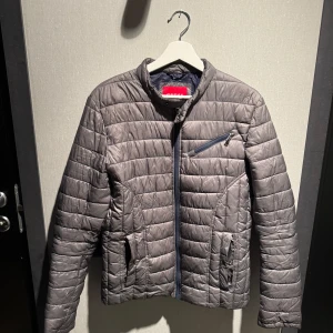 Grå quiltad pufferjacka från Guess - Snygg grå pufferjacka från Guess med quiltad design och dragkedjor på fickorna. Jackan har en hög krage och en bröstficka med dragkedja. Perfekt för kyliga dagar och ger en clean look till din outfit.