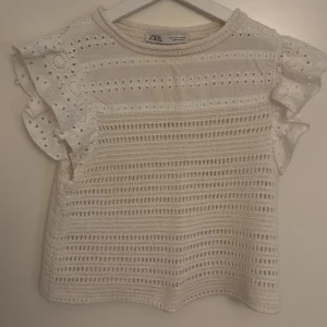 Vit blus med volangärm från Zara - Supersöt vit blus från Zara med broderade detaljer och volangärmar. Toppen har ett luftigt hålmönster och virkade partier som ger en bohemisk vibe. Perfekt till jeans eller kjol för en fräsch look. Rund halsringning och ärmlös med volang.