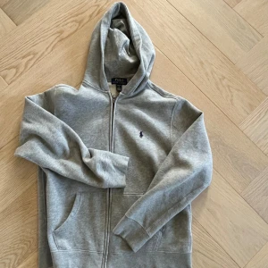 Grå hoodie från Polo Ralph Lauren - Snygg grå hoodie från Polo Ralph Lauren med dragkedja och klassisk broderad logga på bröstet. Tröjan har huva, långa ärmar och två fickor framtill. Perfekt för en chill och avslappnad stil.