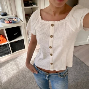 Vit ribbad topp med knappar - Superfin vit ribbad topp med träknappar framtill och fyrkantig halsringning. Modellen är croppad och har korta ärmar som sitter offshoulder. Perfekt till jeans för en fräsch look.