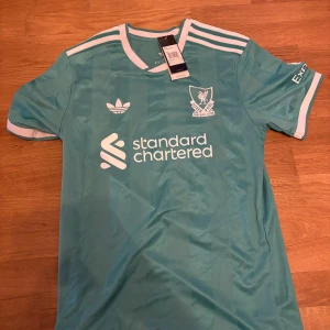 Liverpool Adidas fotbollströja Virgil #4 - Snygg Liverpool fotbollströja från Adidas i ljusblå färg med vita detaljer. Kortärmad modell med klassiska Adidas-ränder på axlarna, Liverpool FC-logga och sponsortryck på bröstet. På ryggen står det 'VIRGIL' och nummer 4. Tillverkad i lätt och ventilerande polyester.