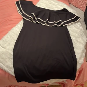 Svart kortklänning med volang - Snygg svart kortklänning med offshoulder och bred volang upptill, kantad med vit detalj. Klänningen har en figurnära passform och är perfekt för dig som gillar en stilren look med lite extra edge. Materialet känns mjukt och stretchigt, vilket gör den bekväm att bära.