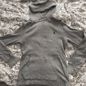 Grå hoodie från Polo Ralph Lauren - Snygg grå hoodie från Polo Ralph Lauren med klassisk broderad logga på bröstet. Tröjan är ribbstickad, har huva med snörning och en stor magficka. Skriv vid fler frågor 