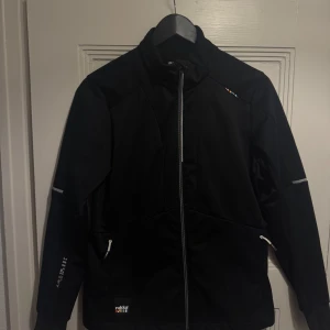 Svart skidjacka från Rukka - storlek 38 - Svart vindjacka från Rukka med hel dragkedja och höga krage. Jackan har reflexdetaljer på ärmarna och små färgglada detaljer vid bröstet och ryggen. Två fickor med dragkedja framtill. Perfekt som skidjacka eller andra utomhusaktiviteter. I mycket gott skick. Storlek 38. 