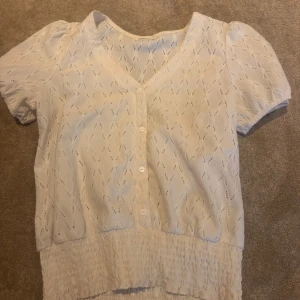 Vit blus med v-ringning och knappar - Supersöt vit blus med v-ringning och korta puffärmar. Blusen har broderat hålmönster, knappar framtill och smockad nederkant som ger en snygg passform. 