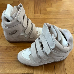 Isabel Marant Skor 38 - Säljer ett par höga sneakers från Isabel Marant i beige mocka med vita detaljer och bruna gummisulor. Skorna har flera breda kardborreband och perforerad vit tå. De har en chunky siluett och coola paneler som ger en unik look.