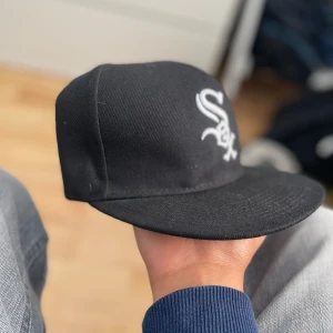 Svart New Era White Sox keps 59FIFTY - Säljer en svart New Era 59FIFTY keps med broderad White Sox-logga i vitt framtill och New Era-logga på sidan. Klassisk basebollkeps med rak skärm och strukturerad passform. Materialet är bomull och polyester, perfekt för dig som gillar sportig streetstyle.