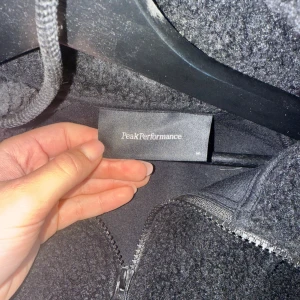 Svart fleece hoodie Peak Performance M - Mysig svart fleece hoodie från Peak Performance med broderad logga på bröstet. Tröjan har huva, lång ärm och en praktisk half zip framtill. Perfekt för kyliga dagar och riktigt skön att dra på sig när det är kallt ute.