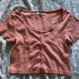 Rosa croppad topp med knappar från lager 157 - Superfin croppad topp i en dämpad rosa färg från lager 157. Modellen har korta ärmar, ribbad struktur och knappar hela vägen framtill. Perfekt till jeans eller kjol för en chill och trendig look. Den är aldrig använd, endast testad. Säljer den eftersom jag aldrig använder den och har en likadan fast i svart. 
