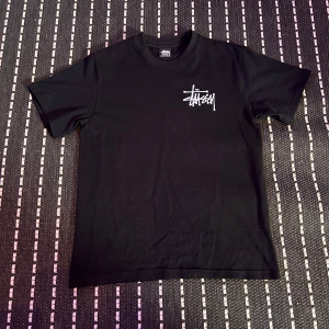 Svart Stüssy t-shirt med logga - Svart t-shirt från Stüssy i bomull med klassisk vit Stüssy-logga tryckt på bröstet och en stor logga på ryggen. T-shirten har rund hals och korta ärmar, perfekt för en streetwear-look.