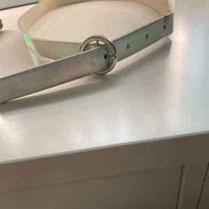 Silvrigt bälte med rund spänne Lindex - Snyggt bälte i silver från Lindex med ett brett band och ett runt metallspänne. Bältet har en cool, glansig finish som ger en futuristisk vibe. Perfekt accessoar för att lyfta din outfit med lite extra edge.