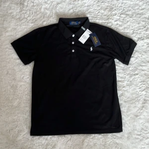 Svart pikétröja från Polo Ralph Lauren - Klassisk svart pikétröja från Polo Ralph Lauren med vit broderad logga på bröstet. Tröjan har korta ärmar, krage och tre knappar framtill. Tillverkad i bomull för en skön och stilren look. Perfekt för dig som gillar tidlös och clean stil.