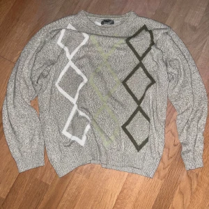 Grå stickad tröja - Säljer en grå stickad tröja från Roger Kent med coolt rutmönster i vitt, grönt och mörkgrönt på framsidan. Tröjan har långärmade ribbade muddar och rund hals. Perfekt för dig som gillar retrovibbar och vill ha något som sticker ut.