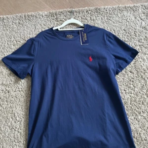 Blå t-shirt från Polo Ralph Lauren - Snygg marinblå t-shirt från Polo Ralph Lauren med klassisk rund hals och korta ärmar. T-shirten är i mjuk bomull och har den ikoniska röda Polo-loggan broderad på bröstet. Perfekt för en chill och stilren look.