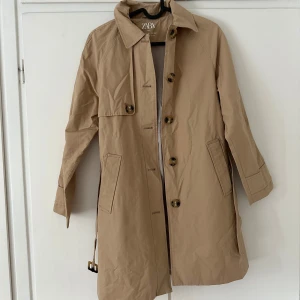 Beige trenchcoat från Zara Kids - Stilren beige trenchcoat från Zara Kids med klassisk krage, knappar framtill och bälte i midjan. Perfekt till hösten! Säljes då den blivit förliten. 