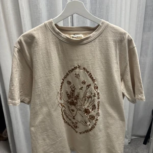 Beige t-shirt med tryck från Acne Studios - Beige t-shirt från Acne Studios med brunt grafiskt tryck framtill som föreställer ett hjorthuvud och blommor. Klassisk rund hals och korta ärmar. Skön och avslappnad passform i mjuk bomull.