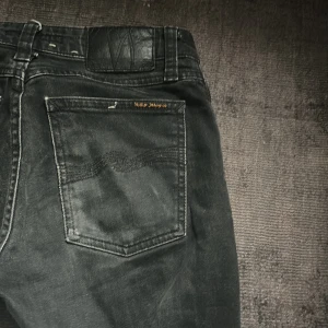 Nudie Jeans - Säljer ett par svarta jeans från Nudie Jeans med klassisk femficksdesign och smal passform. Byxorna har silverfärgade knappar och detaljer samt Nudie Jeans-logga på bakfickan. Perfekta för dig som gillar stilrena och bekväma jeans.