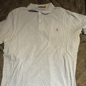 Ljusgrå pikétröja från Ralph Lauren - Säljer en klassisk ljusgrå pikétröja från Ralph Lauren med korta ärmar och krage. Tröjan har två knappar vid halsen och den ikoniska broderade loggan på bröstet. Perfekt för en stilren och avslappnad look. Nya taggen 