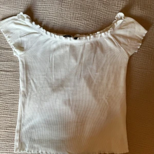 Vit ribbad offshoulder topp från Gina Tricot - Säljer en söt vit ribbad offshoulder topp från Gina Tricot. Toppen har korta ärmar och volangdetaljer längs halsringningen och ärmsluten. 