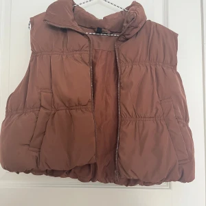 Jacka - Säljer en härlig  brun dunväst från H&M Divided med dragkedja framtill och två fickor. Västen har en hög krage och en puffig, oversized passform och croppad.🥰