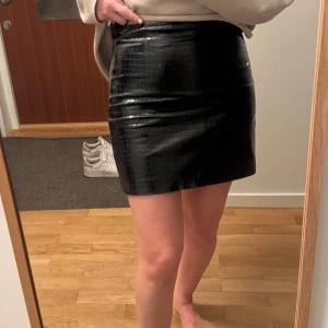 Svart kjol i skinnimitation från Zara - Säljer en svart kort kjol från Zara i skinnimitation med croco-mönster. Kjolen har en tight passform och är perfekt för dig som vill ha en trendig och stilren look. Dragkedja bak för enkel påklädning.