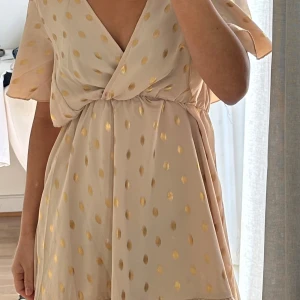 Summer Dress, guldprickar och v-ringning - Säljer en superfin beige kortklänning med glansiga guldprickar och v-ringad omlottfront. Klänningen har korta, vida ärmar och resår i midjan för en bekväm passform. Perfekt för sommarens tillfällen!