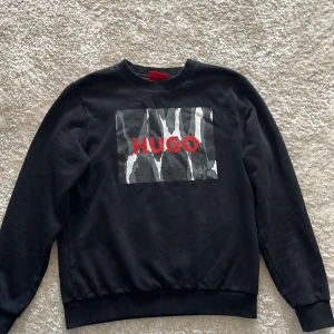 Svart sweatshirt från Hugo med tryck - Svart sweatshirt från Hugo med ett stort grafiskt tryck i grått, vitt och rött framtill där det står 'HUGO'. Tröjan har rund halsringning och ribbade muddar vid ärmslut och nederkant. Perfekt för en stilren och modern look. Rare modell
