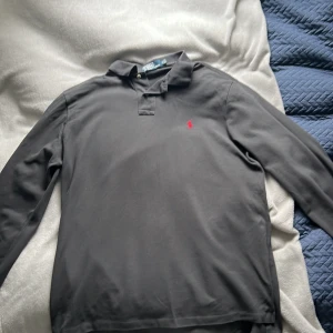 Svart långärmad pikétröja från Polo Ralph Lauren - Klassisk svart långärmad pikétröja från Polo Ralph Lauren med röd broderad logga på bröstet. Tröjan har krage och knäppning med två knappar framtill. Perfekt för en stilren och avslappnad look.