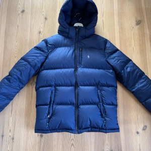 Blå puffjacka från Polo Ralph Lauren - Säljer en blå puffjacka från Polo Ralph Lauren med huva och dragkedja framtill. Nypris över 1700kr