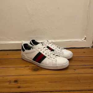 Gucci ace skor - Säljer ett par klassiska vita sneakers från Gucci med gröna och röda ränder på sidorna. Skorna har snörning och rund tå, samt detaljer i grönt på hälen. Perfekta för dig som vill ha en stilren och lyxig look. självklart äkta