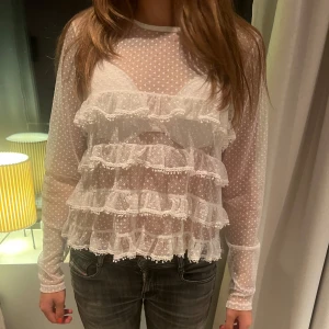 Vit volangblus med prickigt mönster - Säljer en transparent vit blus med prickigt mönster och flera lager volanger framtill. Blusen har långa ärmar och är perfekt för dig som gillar romantiska detaljer och en unik look.