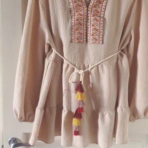 Beige kostym med broderad detalj från SHEIN - Beige kostym från SHEIN med lång ärm och broderad detalj vid halsen. Kostym knytband i midjan med tofsar i rött och gult, samt en lös passform och lätt struktur i tyget.
