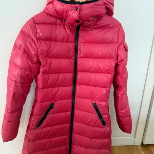 Rosa lång pufferjacka med huva från Moncler - Säljer en snygg rosa pufferjacka från Moncler med huva och svarta dragkedjor. Jackan är lång, har två fickor framtill och quiltad design. Perfekt för kyliga dagar och ger en sportig look. ÄKTA MONCLER köptes för cirka 9000 kr på NK storlek 14 år, men har använts av mig som använder storlek S så passar även en vuxen small. Säljer för 5000 men pris kan diskuteras.