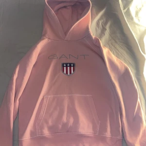 Rosa hoodie från GANT - Säljer en ljusrosa hoodie från GANT med broderad logga på bröstet. Tröjan har huva och en stor magficka framtill. Perfekt för en avslappnad och stilren look. Knappt Använd. Hör av er vid frågor.