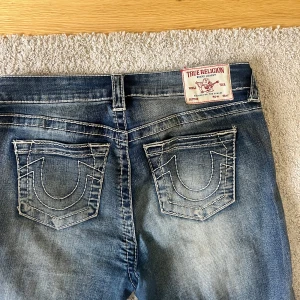 True Religion BECCA Mid Rise Bootcut Jeans - Säljer ett par klassiska blå jeans från True Religion för 400 kr i modell BECCA med midwaist och bootcut. Jeansen har snygga kontrastsömmar och ikoniska bakfickor med broderad detalj. Perfekta för dig som gillar en tidlös jeanslook.
