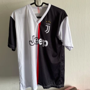 Juventus fotbollströja med Ronaldo 7 - Juventus matchtröja med svart och vit design, röd rand i mitten och Jeep-logga på bröstet. Nummer 7 och Ronaldo på ryggen. Klassisk V-ringning och korta ärmar. 